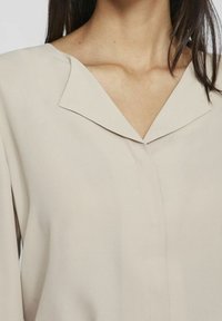 Donna che indossa una blusa beige con un colletto ampio, che mostra il collo e parte dei suoi capelli scuri.
