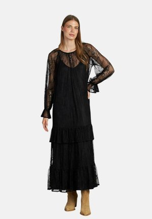 Schwarzes Spitzen-Maxikleid mit langen Ärmeln, Rüscheneinsatz am Saum und fließendem Design. Kombiniert mit sandfarbenen Stiefeletten.