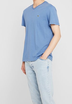 Niebieska koszulka z dekoltem w serek wykonana z miękkiej bawełny, z krótkimi rękawami i małym logo Lacoste na klatce piersiowej. W połączeniu z jasnymi dżinsami.