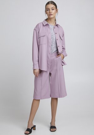 Lila oversized shirt met twee voorzakken, gecombineerd met bijpassende wijde shorts. De outfit wordt compleet gemaakt met zwarte slide sandalen.