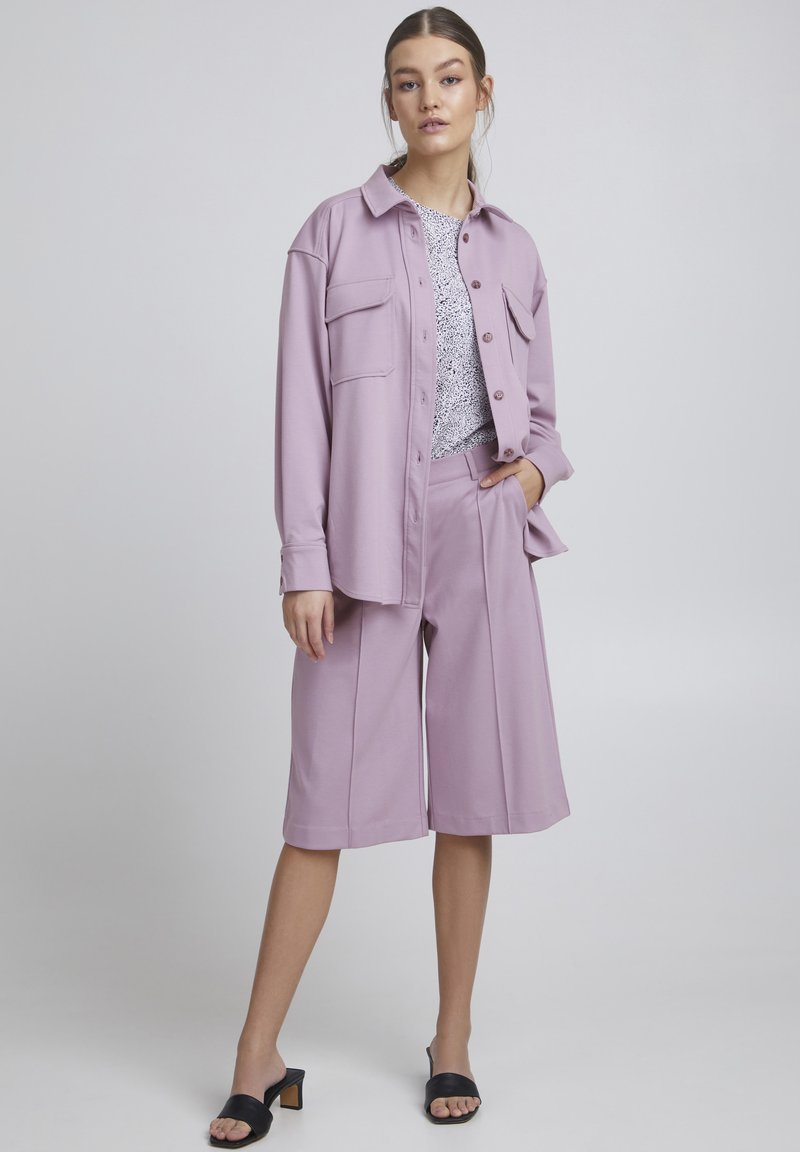 Cămașă oversized de culoare liliac cu două buzunare frontale, asortată cu pantaloni scurți largi în aceeași nuanță.Ținută completată cu sandale negre cu toc plat.
