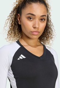 Sort og hvid atletisk top med V-halsdesign. Har Adidas-logo i hvidt på skulderen. Materialet ser glat og let ud.