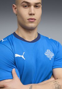 Μπλε αθλητική μπλούζα με αντίθετο σκούρο μπλε γιακά, με λευκά λογότυπα Puma και σχέδιο νιφάδας χιονιού. Μαλακό, υφασμένο ύφασμα.