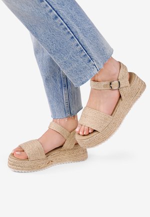 Sandales compensées beige avec texture tressée, bride réglable à la cheville et large bande sur les orteils. Portées avec un jean court bleu clair.