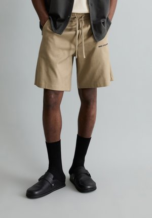 Persoon draagt beige short met trekkoord, zwarte sokken, zwarte instapschoenen en een donker shirt met korte mouwen, met de handen in de zakken.