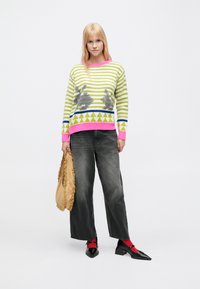 Pletený sveter so zelenými a bielymi pruhmi, ružovými akcentmi, trojuholníkovým vzorom a motívmi zvierat. Kombinovaný so sivými džínsami s širokými nohavicami a čiernymi topánkami.
