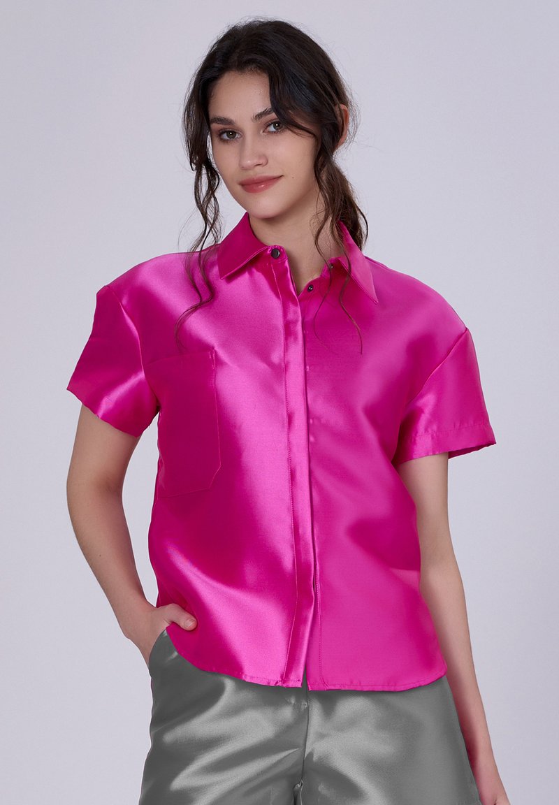 Chemise courte en satin fuchsia à col pointu, avec poche frontale et fermeture à boutons. Texture lisse et finition brillante. Associée à un pantalon en satin gris.