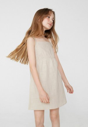 Mladá dívka s dlouhými světlými hnědými vlasy, oblečená v béžových šatech bez rukávů, stojí na jednoduchém bílém pozadí.