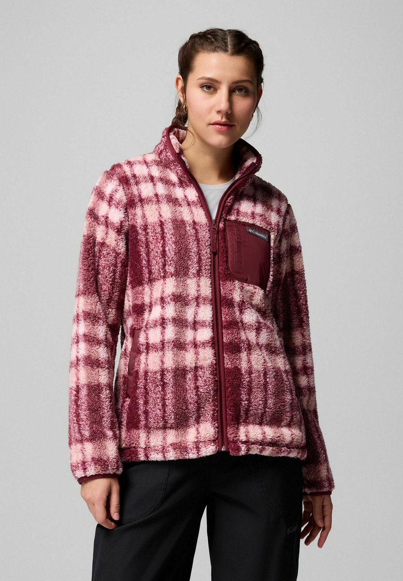 Donna che indossa una giacca in pile a quadri bordeaux e rosa con zip frontale e tasca sul petto, in piedi davanti a uno sfondo grigio uniforme.