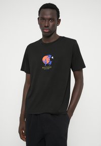 Fekete pólót viselő férfi narancssárga körrel, kék absztrakt madarakkal és sárga "MATISSE ABSTRACT BIRDS" felirattal az elején, fehér háttér előtt áll.