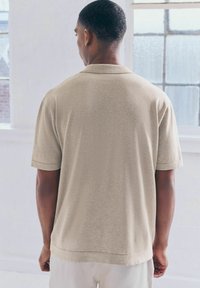 Beige Poloshirt mit kurzen Ärmeln und weicher Textur, mit klassischem Kragen und gerader Saumlinie, von hinten gezeigt.