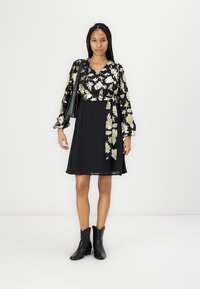 Fekete ruha arany virágos hímzéssel, hosszú ujjakkal és megkötő derékkal; fekete bokacsizmával és fekete kézitáskával párosítva, fehér háttér előtt.