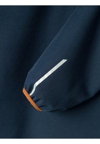 Manga de chaqueta azul marino con puño elástico. Presenta un acento marrón en el puño y una franja diagonal blanca en la manga. Textura suave.