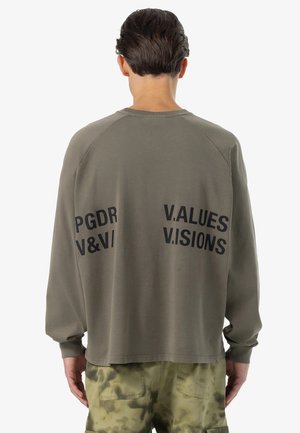 Persoană cu păr scurt și închis la culoare, purtând un hanorac verde măsliniu cu text negru și pantaloni cu model camuflaj, văzută din spate.