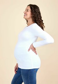Baltoji nėštuminė palaidinė iš raukšlėtos medžiagos, ilgomis rankovėmis ir laisva forma. Dėvinti tamsiai mėlynus džinsus, modelis stovi šonu su matomu nėštumo pilvuku.