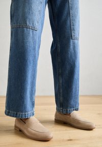 Beige mocka loafers med en texturerad yta, tillsammans med vida blå denimjeans med rå fåll och fickor.