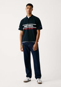 Marinblå kortärmad polo-tröja med vita och röda horisontella ränder och texten "RED BULL RACING", tillsammans med mörk denimjeans och vita sneakers.