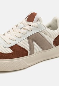 Weißer und brauner Sneaker mit strukturiertem Obermaterial, mit Akzenten aus Leder und Wildleder, runden Schnürsenkeln und einer Gummisohle mit gemustertem Profil.