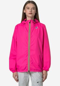 Donna che indossa una giacca con cappuccio rosa acceso con cerniera a righe multicolori e pantaloni grigio chiaro, in piedi su sfondo bianco.