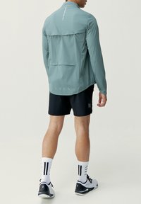 Lätt, mintgröna jacka med hög krage, bakventil och reflekterande detaljer, tillsammans med svarta shorts och mönstrade strumpor.