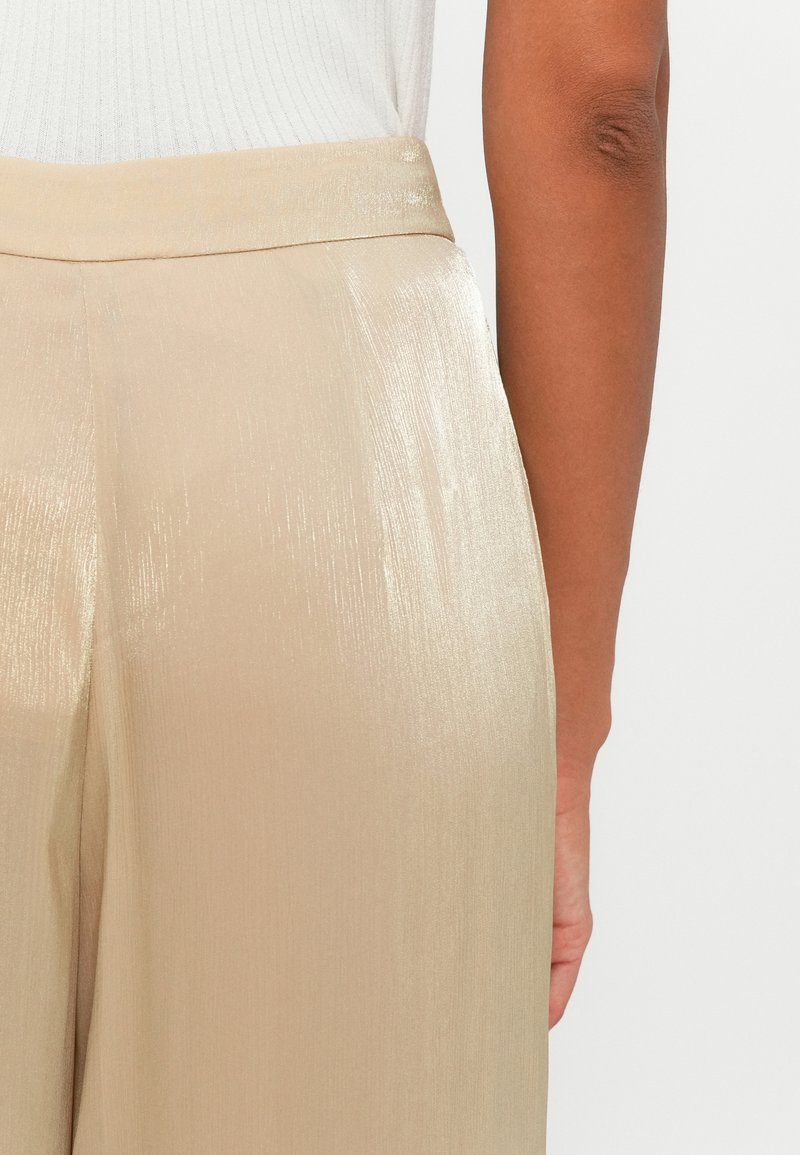 Beige, hochgeschnittene Hose mit metallischem Glanz, die eine glatte Textur und dezente vertikale Streifen aufweist, von hinten gezeigt.