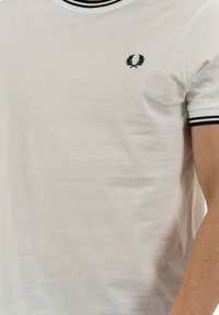 Camiseta blanca de manga corta hecha de algodón, con cuello y bordes de las mangas en canalé negro, y un pequeño emblema bordado en negro en el pecho.