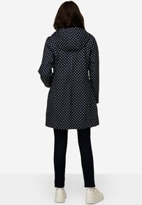 Laivastonsininen pilkkusateenpitävä takki hupulla, keskipitkä malli ja vetoketjukiinnitys, yhdistettynä tummiin farkkuihin ja vaaleisiin jalkineisiin.