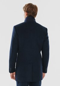Granatowy wełniany płaszcz o dopasowanym kroju, z wysokim kołnierzem, mankietami na cztery guziki oraz rozcięciem z tyłu, charakteryzujący się gładką fakturą.
