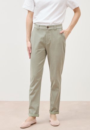 Osoba ubrana w podwinięte jasne oliwkowe spodnie, białą koszulę z krótkim rękawem oraz różowe teksturowane buty typu slip-on, stojąca na neutralnej podłodze.