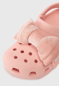 Clog rosa perfurado com um grande laço de tecido macio na parte superior, apresentando sola texturizada e tira traseira ajustável.