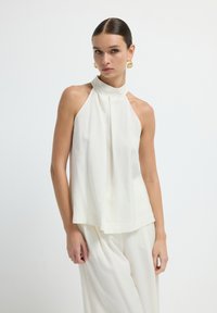 Top halter blanc sans manches avec un col haut, des détails plissés et une coupe fluide. Texture de tissu lisse, design minimaliste.