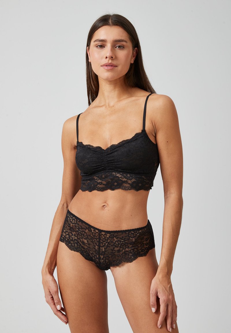 Bralette en dentelle noire avec des bords festonnés et des bretelles réglables, associé à un short en dentelle à taille basse assorti présentant un design festonné similaire.
