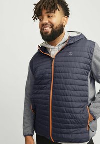 Mørkeblå isoleret vest med vandret quiltet mønster, orange lynlås og hætte; grå langærmet skjorte under; to sidelommer.