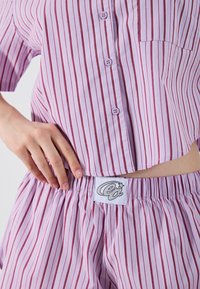 Ensemble de pyjama violet clair avec des rayures verticales bordeaux, comprenant un haut à boutons et un short avec taille élastique, incluant une étiquette de marque sur le short.