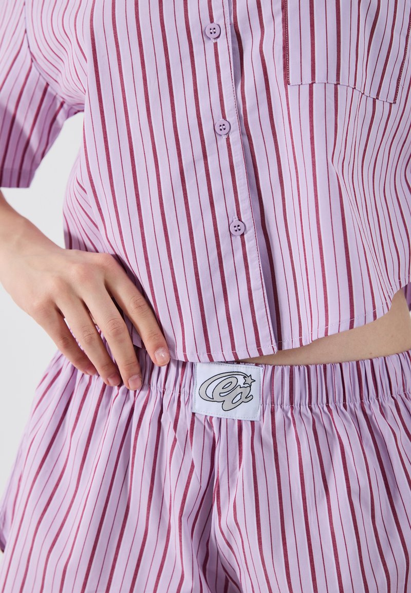 Pigiama di colore lilla chiaro con strisce verticali bordeaux, composto da una maglietta con bottoni e pantaloni corti con vita elasticizzata, include un'etichetta di marca sui pantaloni.