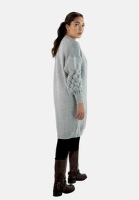 Übergroßer grauer Strickpullover mit strukturierten Ärmeln im Bubbelmuster, kombiniert mit schwarzen Leggings und braunen knielangen Stiefeln.