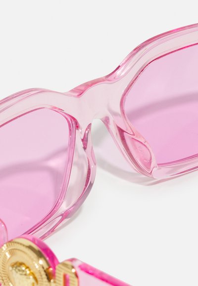 Óculos de sol rosa com uma armação transparente rosa e lentes graduadas em rosa. Apresenta detalhes em hardware dourado nas hastes. Textura lisa e brilhante.