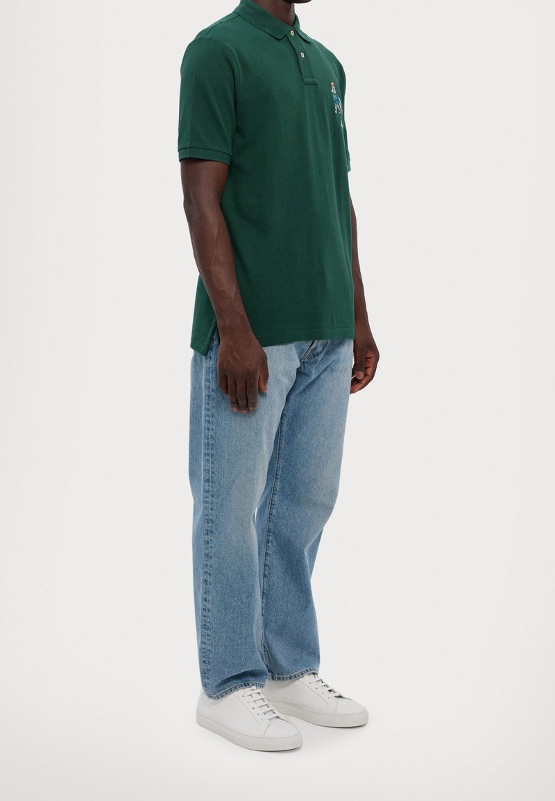 Homme portant un polo vert foncé, un jean droit bleu clair et des baskets blanches, se tenant contre un fond blanc uni.