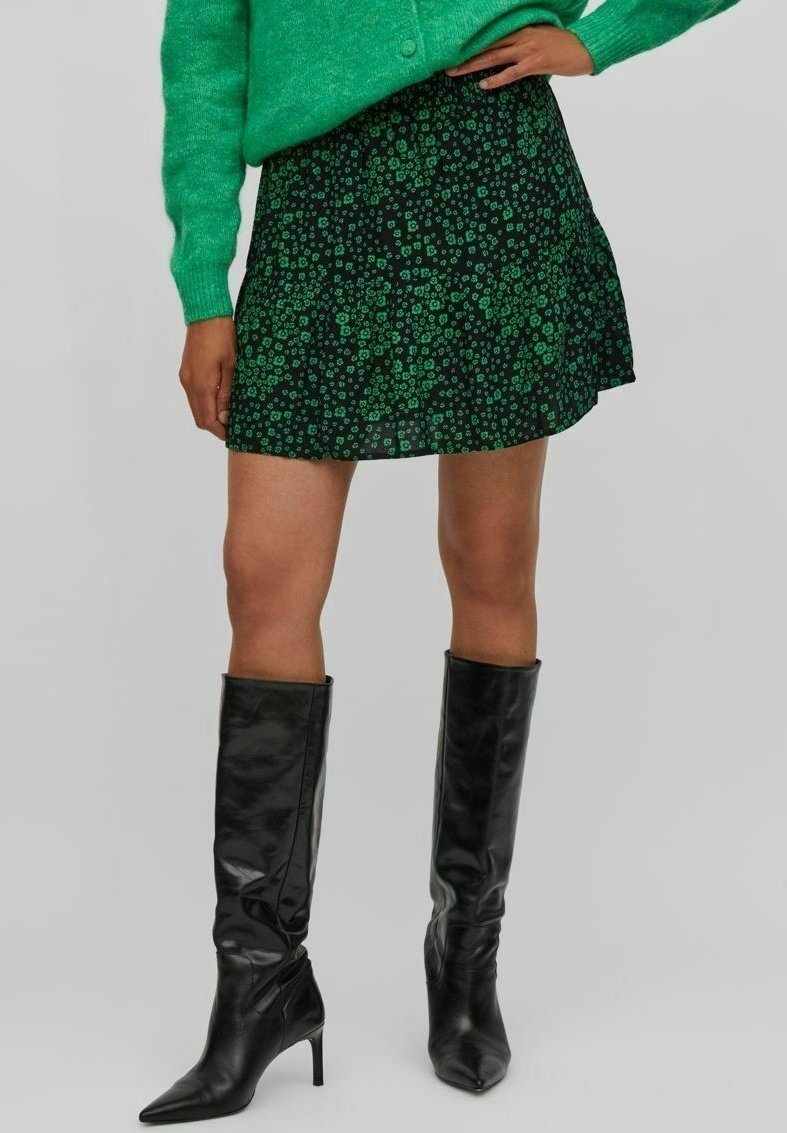 Jupe verte à motif floral avec ourlet évasé, associée à des bottes noires en cuir à talons aiguilles montant jusqu'aux genoux, avec une finition lisse et un bout pointu.