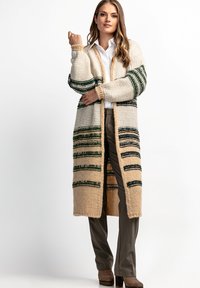 Sweter z dzianiny w kolorze kremowym, z zielonymi paskami i beżowymi akcentami, charakteryzujący się długim, otwartym krojem oraz ściągaczami na rękawach. Połączony z szarymi spodniami.