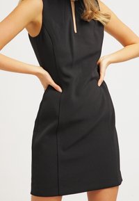 Robe noire sans manches avec un col haut et une découpe en œil-de-bœuf, coupe ajustée, texture lisse et coutures structurées.