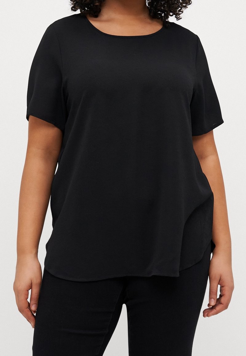 Femme portant un haut noir uni à manches courtes et un pantalon noir, debout les bras détendus le long du corps devant un fond uni.