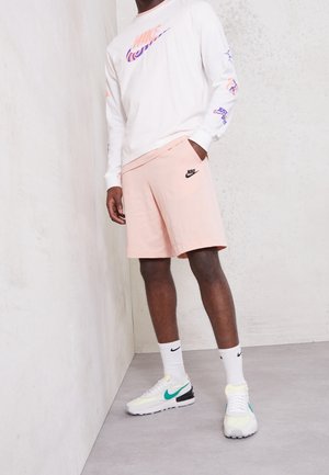 Camisa blanca de manga larga con logo de Nike en púrpura, combinada con pantalones cortos rosas con logo de Nike en negro, calcetas blancas y zapatillas con detalles en verde.