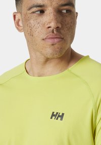 Žluté atletické tričko má texturovanou tkaninu, kulatý výstřih a vyšívané černé logo na hrudi. Pohled zblízka.