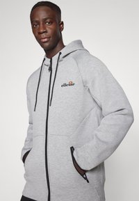 Sivi zip-up dukserica s crnim šavovima, elastičnim vezicama i džepovima. Ima mali izvezeni logo na grudima. Mekana tekstura.