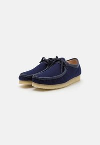 Sapatos em feltro azul marinho com detalhes em couro liso, apresentando fecho com atacadores e uma sola em borracha bege clara para contraste.