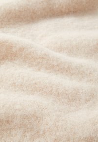 Tissu beige clair avec une texture douce et floue, présentant un motif de surface subtil et irrégulier ainsi que des plis délicats. Idéal pour des vêtements chauds ou des couvertures.