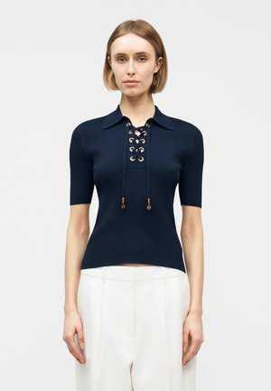 Frau trägt ein marineblaues, geripptes Kurzarmoberteil mit Schnürung vorne und weiße, hoch taillierte Hosen, steht vor einem einfarbigen Hintergrund.