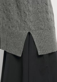 Pull gris tricoté avec un motif torsadé, ourlet côtelé et fentes latérales. Porté par-dessus un vêtement noir, mettant en valeur la texture et les superpositions.