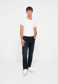 Jeans oscuros de hombre en corte recto, combinados con una camiseta blanca ajustada. Zapatillas blancas con acentos grises completan el look.
